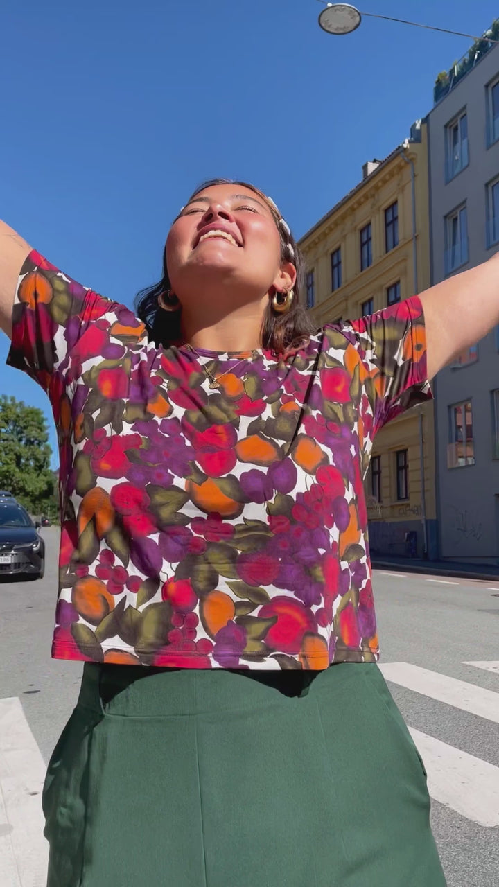 T-skjorte med frukt print fra Dianas Vintage. Hvit bunn med lilla, orange og grønn frukt. Rund hals, korte ermer og kort lengde.