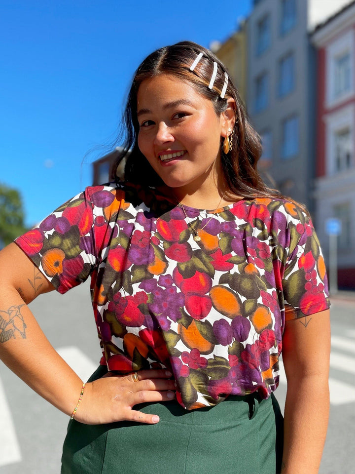 T-skjorte med frukt print fra Dianas Vintage. Hvit bunn med lilla, orange og grønn frukt. Rund hals, korte ermer og kort lengde.